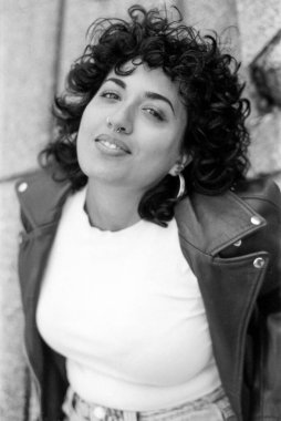 Tahıllı monokrom analog görüntüsü kıvırcık saçlı, burun piercingli, halka küpeli, beyaz tişörtlü, deri ceketli, havalı ve kendinden emin tavırları olan bir kadın.