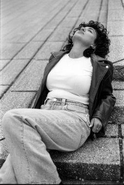Tahıllı, tek renkli, kıvırcık saçlı, deri ceket ve kot giymiş, kentsel bir ortamda beton basamaklarda gevşeyen genç bir kadını çeken bir fotoğraf.