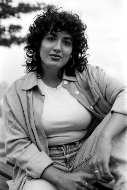 Koyu renkli analog bir görüntü, kıvırcık saçlı, burun halkalı, açık renk ceketli, tişörtlü ve kot pantolonlu, açık havada, düşünceli bir ifadeyle oturan genç bir kadını çekiyor.