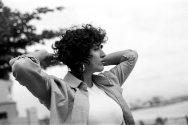 Siyah-beyaz, kıvırcık saçlı, beyaz bir tişörtün üzerine açık keten gömlek giyen, saçını tutan ve kameradan gözünü ayırmayan genç bir kadının görüntüsü.