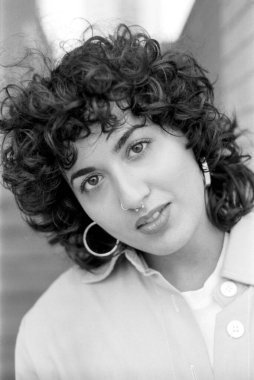 Tahıllı monokrom analog fotoğraf kıvırcık saçlı, burun piercingli, geniş halka küpeli, beyaz bir gömleğin üzerine açık renk ceket giyen, klasik bir estetik yaratan genç bir kadını çekiyor.