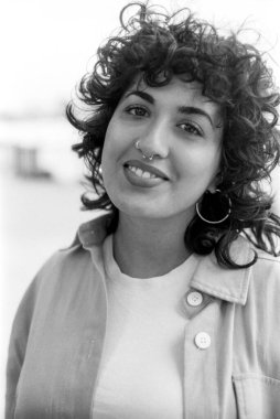 Tahıllı monokrom analog fotoğraf kıvırcık saçlı, burun halkalı, halka küpeli, beyaz bir tişörtün üzerinde rahat bir şekilde gülümseyen genç bir kadını çekiyor.