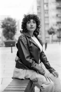 Tahıllı, tek renkli, kıvırcık saçlı, burnu delinmiş, deri ceketli, halka küpeli, şehir merkezinde düşük tuğlalı bir duvarda oturan genç bir kadının görüntüsü.