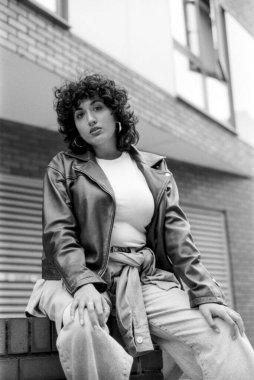 Tahıllı, tek renkli, analog tarzda, kıvırcık saçlı ve deri ceketli genç bir kadının şehir ortamında serin ve kendinden emin bir tavır sergilemesi.