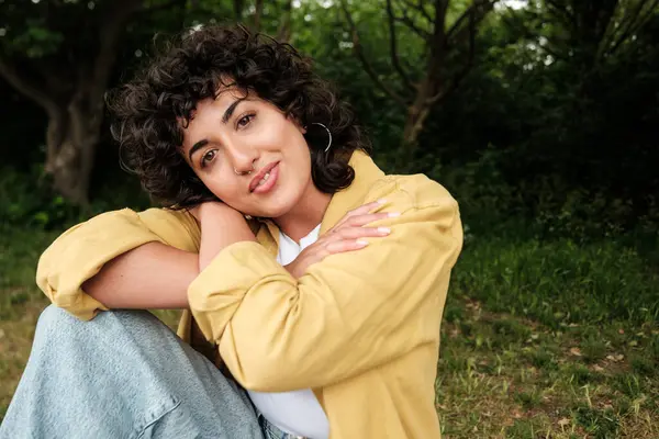 Huzurlu, doğal bir ortamda oturan genç bir kadın, nazik bir gülümseme, kendine güven, kendini kabullenme ve doğayla derin bir bağ ile kendini kucaklıyor.