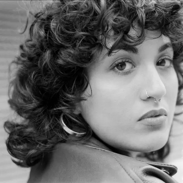 Tahıllı monokrom analog fotoğraf kıvırcık saçlı, burun piercingli, büyük halka küpeli deri ceket giyen ve kendine güveni tam bir kadın yakalamış.