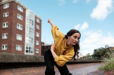 Yoğunlaşmış genç dansçı kadın çatıda modern bir dans koreografisi uyguluyor. Arka planda tuğla bina ve bulutlu gökyüzü var. Özgürlük ve şehir tarzı ifade ediyor.