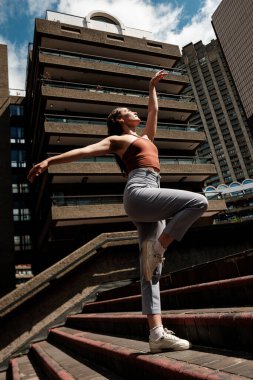 Genç bir kadın Londra 'daki Barbican malikanesinin merdivenlerinde zarif bir şekilde dans ediyor. Güneşli bir günde vahşi mimarinin arka planına karşı yeteneğini sergiliyor.