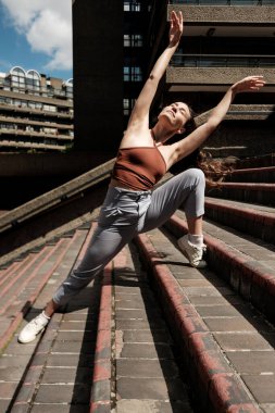 Genç bayan dansçı şehir merkezindeki bir merdivende modern bir dans gösterisi sergiliyor, çarpıcı bir mimarinin arka planına karşı akıcılık ve zarafet ifade ediyor.