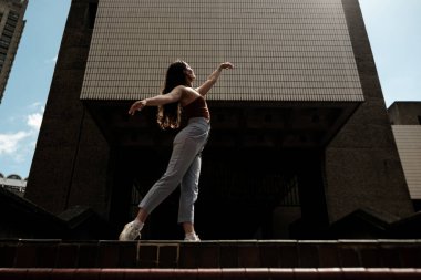 Genç bir kadın şehir ortamında zarif bir şekilde dans ediyor, kolları açık ve vücudu modern mimarinin arka planına karşı hareketin neşesini ifade ediyor.