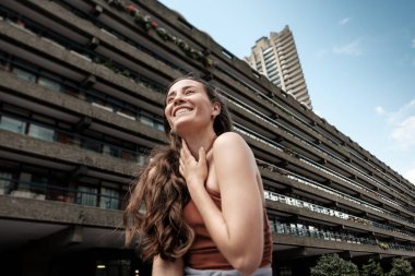 Uzun kahverengi saçlı mutlu bir kadın boynuna dokunuyor ve gülümsüyor. Güneşli bir günde Londra, İngiltere 'de, Barbika Malikanesi ve Trellick Kulesi' nin önünde dururken.