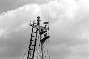 Üzerinde merdiven, navigasyon ışıkları, anten ve bulutlu bir gökyüzüne karşı kurulmuş diğer ekipmanların yer aldığı bir gemi direği tasvir edilen taneli monokrom analog klasik fotoğraf.