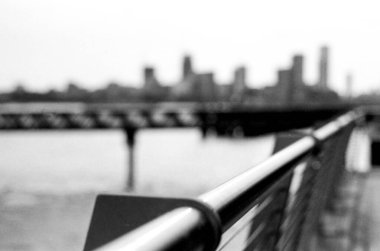 Taneli tek renkli analog fotoğraf keskin odak noktasında metal bir korkuluk yakalıyor, bulanık bir şehir manzarasına doğru gidiyor, bir mesafe ve şehir atmosferi hissi yaratıyor.