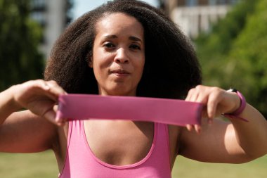 Pembe bir direniş bandı tutan kararlı bir kadın, canlı bir açık hava parkında spor rutinine konsantre oluyor, yemyeşil ve sağlıklı, aktif bir hayat sürüyor.