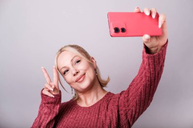 Gülümseyen bir etken, parlak gri arka planda akıllı bir telefonla selfie çekerken barış işareti yapıyor, modern dijital yaşam tarzı ve sosyal medya bağlantısını somutlaştırıyor.