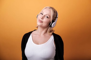 Beyaz kulaklık takan genç sarışın kadın müziği seviyor, gözleri kapalı ve turuncu arka planda, canlı ve neşeli bir atmosfer yaratıyor.