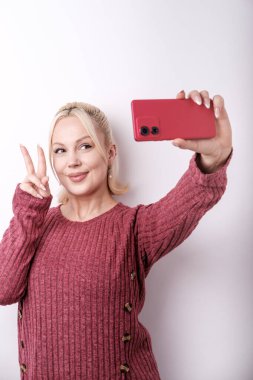 Genç nüfuzlu biri gülümseyerek zafer işareti yapıyor ve kırmızı akıllı telefonuyla selfie çekiyor. Günlük kıyafetler giyip mutluluk saçıyor.