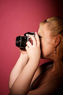 Genç fotoğrafçı klasik bir analojik kamera kullanarak fotoğraf çekiyor, pembe arka planda dikkatle odaklanıyor, fotoğrafçılık ve retro estetik tutkusunu sergiliyor.