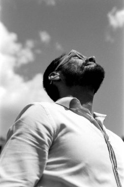 Klasik, kumlu, siyah ve beyaz 35 mm 'lik analog fotoğraf, gözlüklü bir adamı bulutlu bir gökyüzüne bakarken yakalar, düşünceli ve atmosferik bir sahne yaratır.