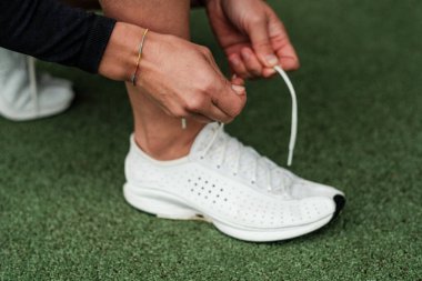 Beyaz koşu ayakkabılarının bağcıklarını bağlamaya odaklanmış bir sporcunun yakın plan görüntüsü canlı yeşil bir çimenlikte yoğun bir antrenmana ya da antrenmana hazırlanıyor.