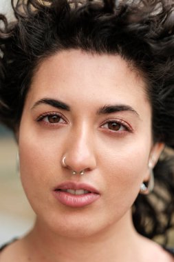 Yakından çekilmiş bir portre kıvırcık saçlı, burnu delinmiş, kahverengi gözlü, kendine güvenen ve benzersiz kişiliğini sergileyen çarpıcı bir genç kadın.