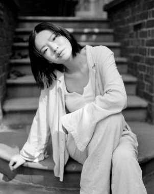 Çekici monokrom orta formatlı film fotoğrafında kısa saçlı şık Asyalı bir kadın açık hava merdivenlerinde açık renkli kıyafetlerle oturuyor, gelişigüzel şehir zarafeti yayıyor.