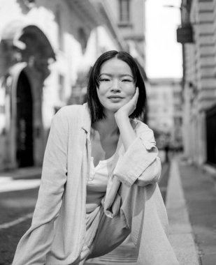 Kentte çömelmiş genç bir Asyalı kadının tek renkli orta formatlı bir film portresi. Çenesini eline dayayıp dinleniyor ve günlük giysiler giyiyor.