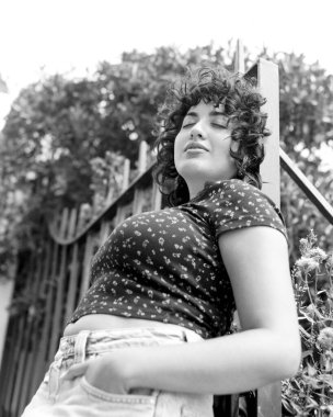 Siyah ve beyaz, kıvırcık saçlı, gözleri kapalı, metal korkuluklara yaslanmış, sıcak bir yaz gününün tadını çıkaran, kumlu, tek renkli estetik bir kadının orta sınıf bir film portresi.