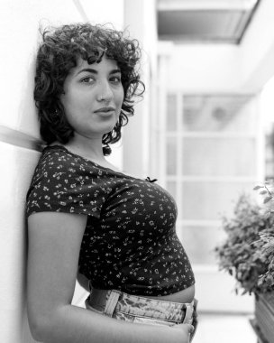 Tek renkli orta formatlı film portresi kıvırcık saçlı ve burun halkalı kendine güvenen bir genç kadın, sıradan kıyafetlerle duvara yaslanmış, kişisel bir stil yayıyor.