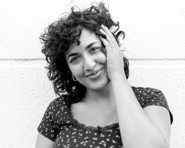 Tek renkli orta formatlı, kıvırcık saçlı, burun piercingli, yüzüne hafifçe dokunan ve beyaz bir duvarın yanında gülümseyen neşeli genç bir kadının portresi.