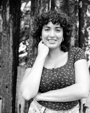 Orta formatta, kıvırcık saçlı, burnu delinmiş, eli çenesinde, eskimiş ahşap bir çitin önünde duran, kendine güveni ve doğal güzelliği olan neşeli bir genç kadının portresi.