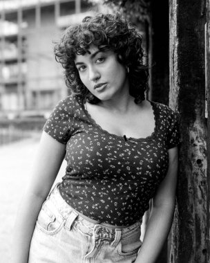 Tek renkli orta formatta, kıvırcık saçlı ve burnu piercingli, şehir ortamında eskimiş ahşap bir direğe yaslanmış şık bir genç kadının film portresi.