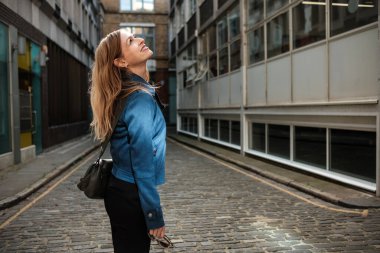 Mavi ceket giyen ve kurye çantası taşıyan profesyonel bir kadın Londra 'daki dar bir kaldırım taşından sokak aralığındaki binaların mimarisine hayran kalıyor.