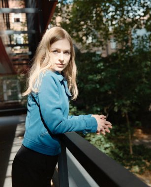 Mavi ceketli ve siyah pantolonlu sarışın kadın korkuluklara yaslanıyor, ağaçlarla güneşli bir şehir gününün tadını çıkarıyor ve klasik, kumlu bir film tarzında bir yürüyüş yolu izliyor.