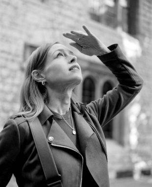 Kasvetli monokrom orta formatlı film portresi. Şehir mimarisinin ortasında merak ve keşif duygusunu somutlaştıran hareketli bir şehirde gözlerini yukardan yukarıya dikmiş bir kadının portresi.