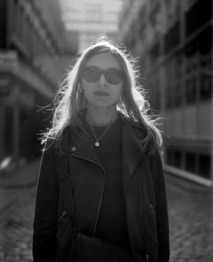 Güneş gözlüğü ve deri ceket takan şık bir genç kadının tek renkli orta formatlı film portresi. Dar bir şehir sokağında saçlarını aydınlatan güneş ışığıyla duruyor.
