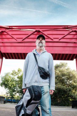 Gri kapüşonlu, beysbol şapkalı ve seyahat çantalı yaşlı evsiz bir adamın portresi kırmızı bir köprünün altında dikilip yukarı bakıyor, şehirde evsizlik ve yoksulluk yaşıyor.