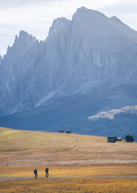 Sonbahar manzarasında dev kaya dağları ve kulübelerin önünde yürüyen iki yürüyüşçü, dikey, Dolomitler..
