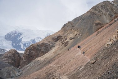 Buzulları ve arka planda karlı zirveleri olan kayalık arazide yürüyüş yapan turistler, Pamir, Kırgızistan.