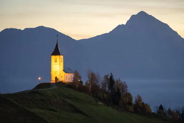 Gün doğarken, Slovenya ve AB 'de dağlarla çevrili bir tepenin üzerinde küçük aydınlatılmış bir kilise.