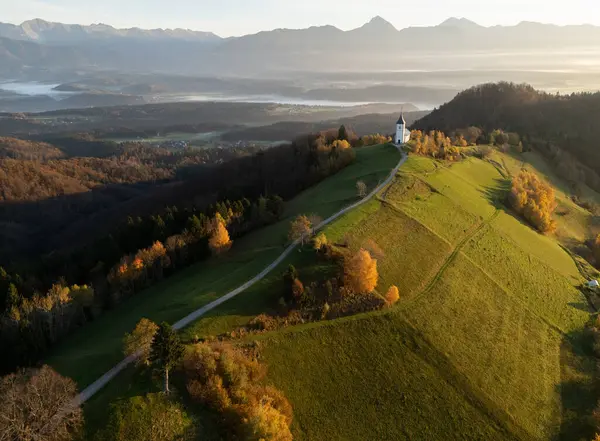Slovenya 'da bir tepenin tepesinde küçük bir kilise ve arka planda dağlar bulunan havadan dağlık bir arazi..