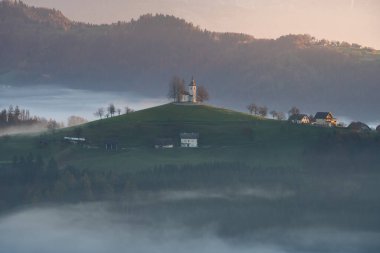 Slovenya ve Avrupa 'nın renkli gündoğumu sırasında etrafı sislerle çevrili bir dağın tepesindeki küçük kilise.