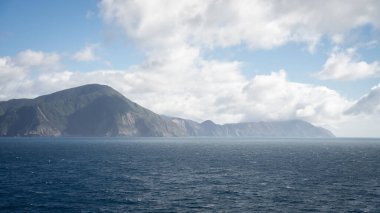 Bulutlu bir günde yelken açan tekneden dağlık sahile bakın, Yeni Zelanda.