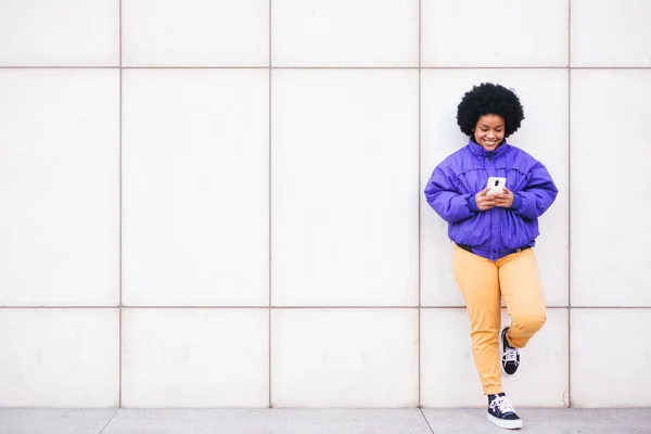 young latin afro woman standing on white wall using phone and smiling, space for copy. High quality photo