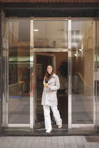 Confident young woman going out for a walk, latin ethnicity. She is leaving the portal building holding a yellow cellphone