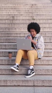 Laptopta güzel haberleri kutlayan heyecanlı afro kadın, şehir merdivenlerinde oturdu. Yüksek kalite fotoğraf