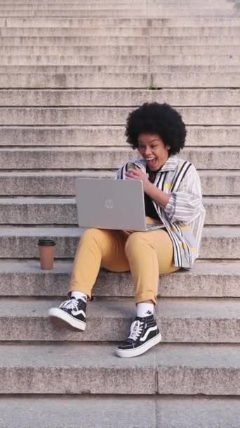 兴奋的非洲女人坐在城市的楼梯上 一边用笔记本电脑庆祝好消息 高质量的照片 — 图库视频影像