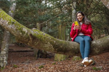 Ormanda cep telefonuyla ağaç dalında oturan neşeli kadın. Yüksek kalite fotoğraf