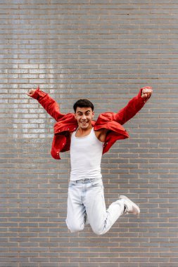 Latin LGBT young man with red overshirt jumping with joy in front of a brick wall clipart