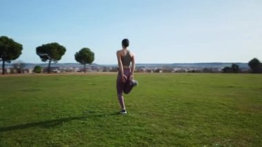 Spor kıyafetleri giyen genç bir kadın yeşil bir parkta bacak esneme gösterisi yapıyor.